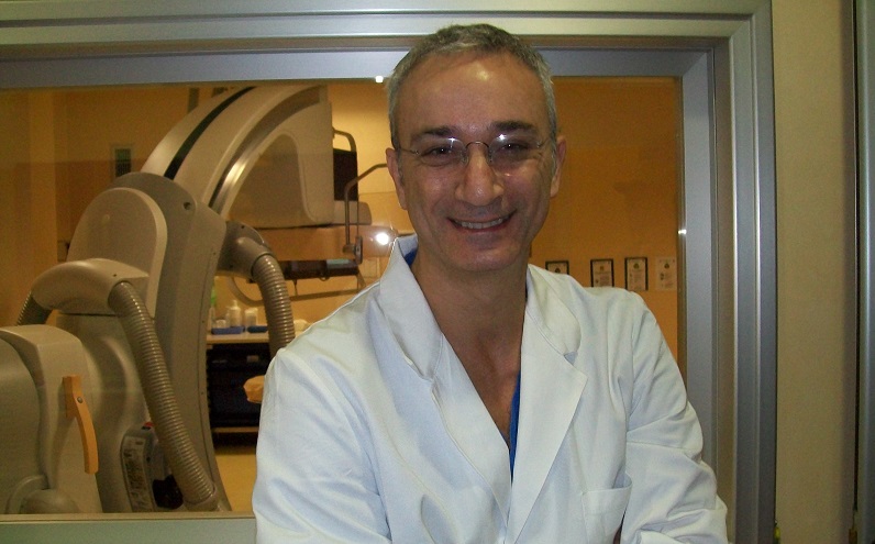 Nella foto il dottor Marco Comeglio, con il camice, è visibile solo fino alle braccia, in secondo piano si vede un'apertura con un vetro e dietro una parte della sala di emodinamica
