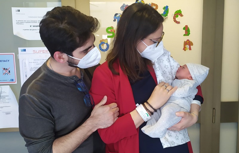 Nella foto si vedono i neogenitori in piedi, davanti alla porta del reparto. La neonata è in braccio alla mamma e il babbo ha un braccio sopra la spella della mamma